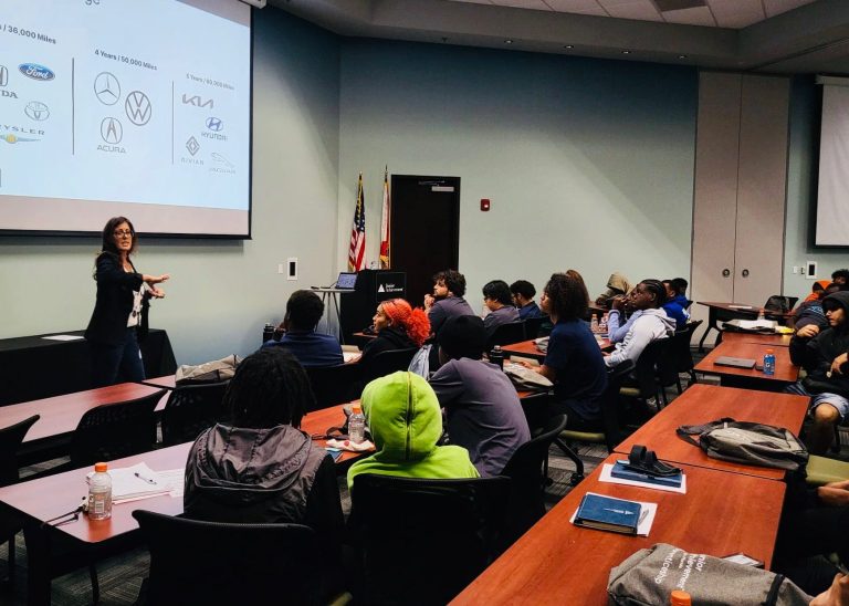 A presenter speaks to an audience in a classroom setting with visual aids on the screen.